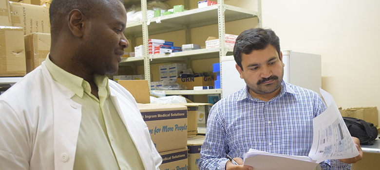 Two men stand in a pharmacy storeroom looking over papers.