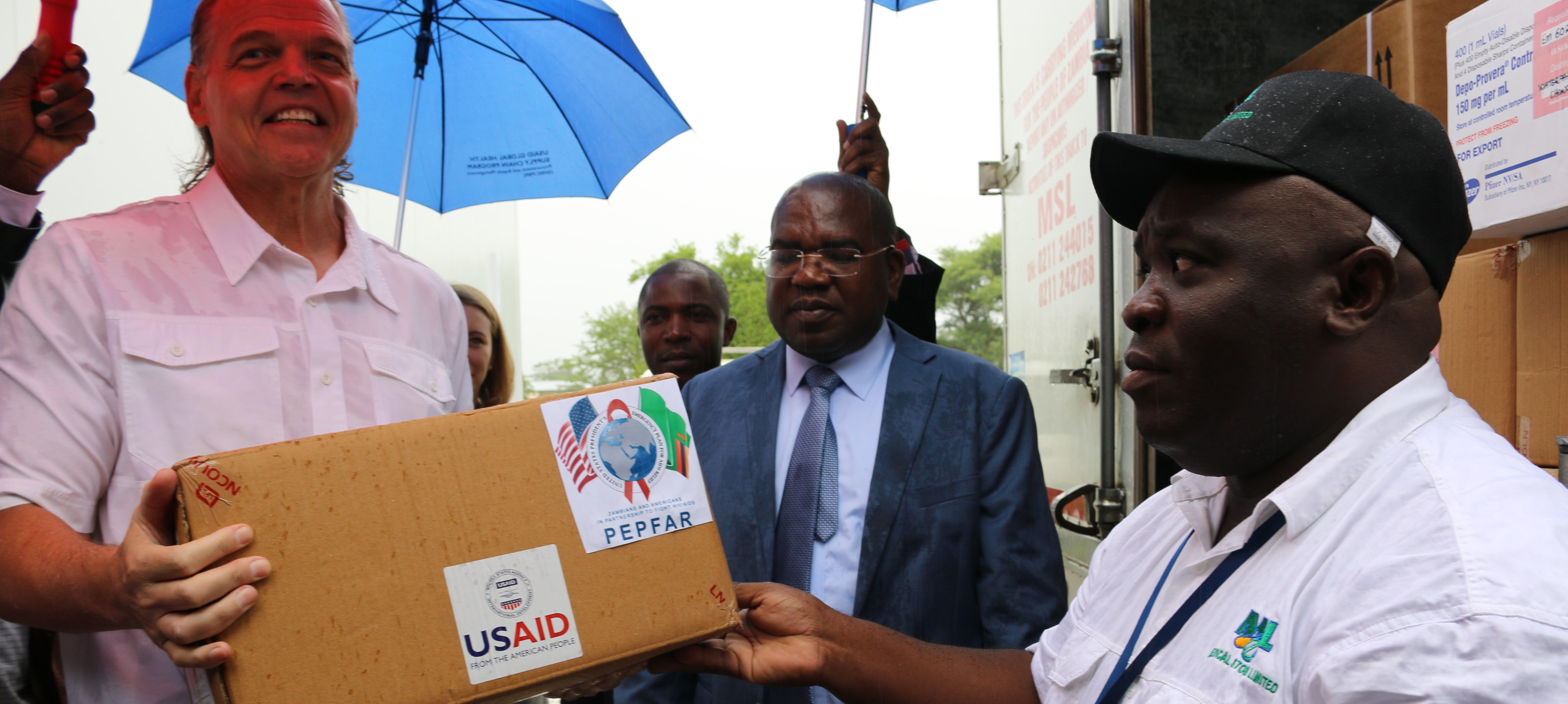 Ambassador Daniel Foote offloads a box of health commodities to be moved into a pre-fabricated storage unit as Zambian Minister of Health Chilufya and MSL Managing Director Chikuta Mbewe look on.