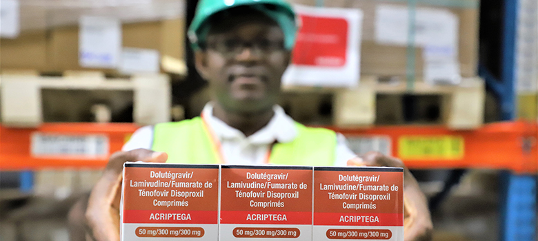 A man wearing a yellow vest and green safety helmet in a warehouse holds up a box of anitretrovirals.