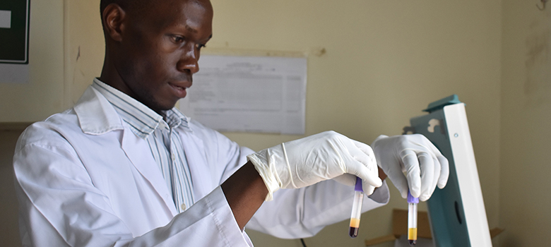 A Ghanaian man in a striped button-up shirt and lab coat holds up two test tubes.