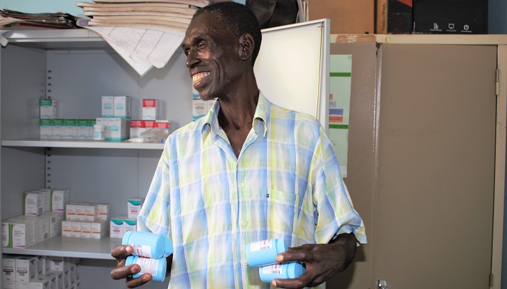Image of Jolly Mubanga HIV patient in a clinic in Zambia holding several bottles of TLD and smiling. 
