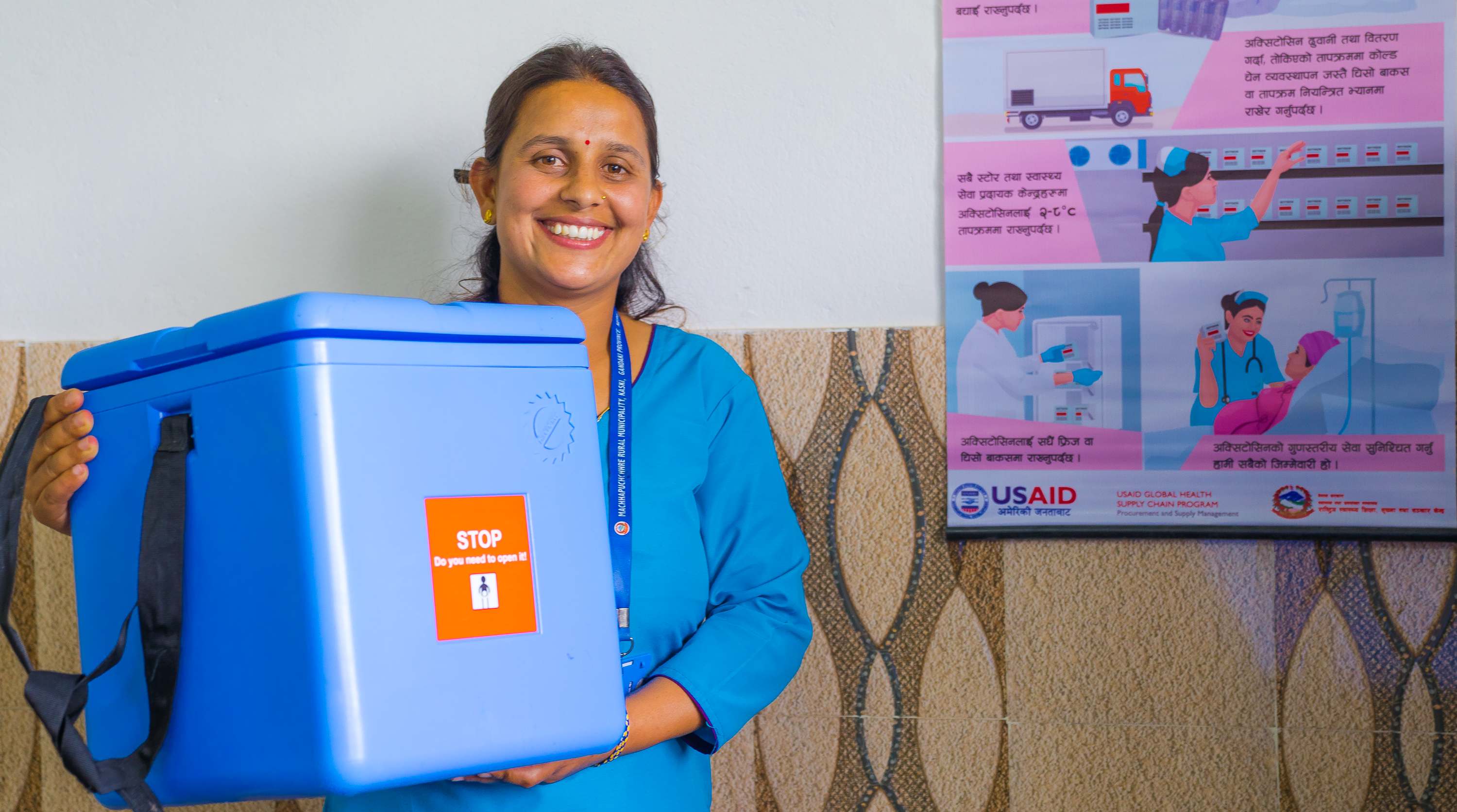 Nepali nurse carries cold box for health supplies near poster about proper oxytocin storage.