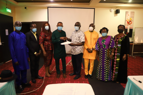 A group of Nigerian government representatives stand in a semicircle and hold a paper agreement, two in center shake hands