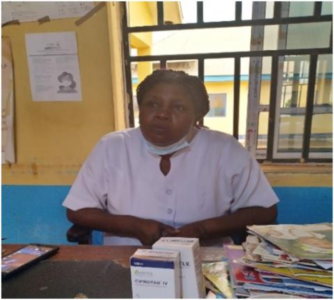 Nigerian health worker sits at desk with blue and yellow wall and window in background, medicines and papers on desk